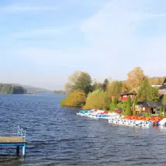Widok na jezioro z pomostem i kolorowymi rowerami wodnymi przy brzegu, w tle domy i jesienne drzewa.