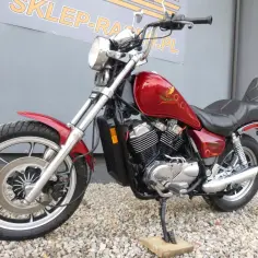 Czerwony motocykl Honda Shadow 500 z chromowanymi elementami, zaparkowany na żwirze przed szarym budynkiem.