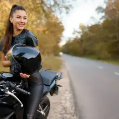 Uśmiechnięta kobieta w skórzanej kurtce i legginsach trzyma kask obok czarnego motocykla. W tle droga i jesienne drzewa, idealne do testowania różnych marki motocykli.