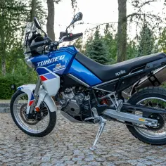 Błękitny motocykl Aprilia Tuareg gotowy na przygodę, zaparkowany na kamiennej nawierzchni wśród drzew.