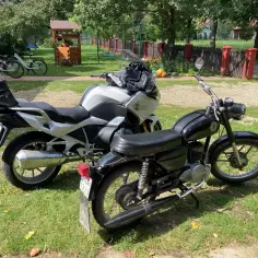 Dwa motocykle, jeden nowoczesny srebrny, drugi klasyczny czarny, stoją na trawie. Obok rower.