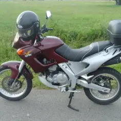 Ciemnoczerwony motocykl Aprilia Pegaso 650 z czarnym kufrem i kaskiem, gotowy na przygodę na tle zielonego pola.