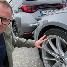 Mężczyzna w okularach pokazuje felgę Audi. Za nim szare auto z niemiecką tablicą. Szukasz tanich motocykli z Niemiec?