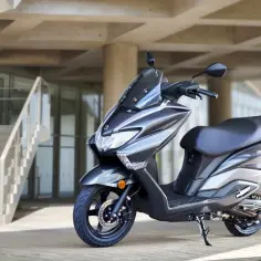 Szary skuter Suzuki Burgman 125, jeden z najbardziej bezawaryjnych skuterów 125, stoi przed nowoczesnym budynkiem z betonowymi elementami i schodami.