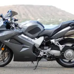 Szary motocykl Honda VFR 800 z pełnym owiewkami, zaparkowany na tle krajobrazu z wodą i wzgórzem.