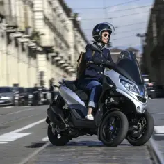Kobieta w kasku i okularach prowadzi srebrny motocykl trójkołowy na kat B, przemierzając ulicę miasta.
