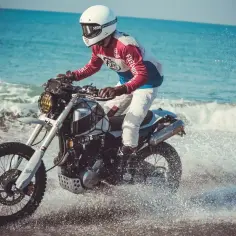 Motocyklista w kasku jedzie motocyklem po plaży, rozpryskując wodę.