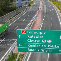 Zielona ciężarówka z piaskiem jedzie drogą krajową 91. Znak kieruje do Podwarpia, Katowic, Cieszyna, Kamienicy Polskiej i Rudnika Wielkiego.