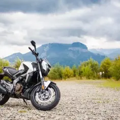 Biało-czarny Bajaj Dominar 400 na kamienistym podjeździe, w tle góry i zielone drzewa pod pochmurnym niebem.
