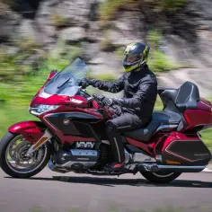 Mężczyzna w kasku jedzie lśniącym, czerwonym motocyklem honda turystyk. W tle rozmyty krajobraz z zielenią i skałami.