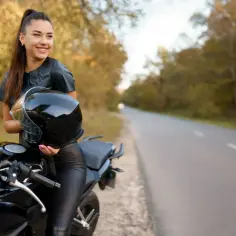 Uśmiechnięta kobieta w skórzanym stroju trzyma kask obok swojego czarnego motocykla. Idealny motocykl dla kobiety na jesienną przejażdżkę.