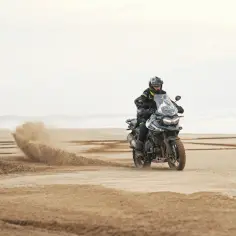 Motocyklista w kasku i kurtce przemierza piaszczystą plażę, zostawiając za sobą chmurę piasku. To prawdziwa przygoda na drodze szutrowej.