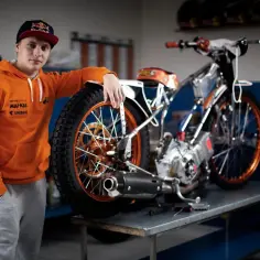 Młody żużlowiec w pomarańczowej bluzie opiera się o swój motocykl żużlowy, gotowy do wyścigu.