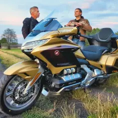 Złoty Honda Goldwing na tle wiejskiego krajobrazu. Dwóch mężczyzn rozmawia obok potężnej maszyny.