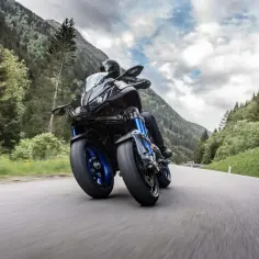 Motocyklista na Yamaha Niken pokonuje zakręt na górskiej drodze, otoczony zielenią i chmurami.