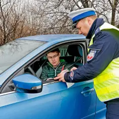 Policjant wręcza kierowcy mandat. Młody mężczyzna w okularach, siedzący za kierownicą niebieskiego auta, przyjmuje dokument. Czy to jego recydywa mandat?