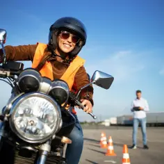 Uśmiechnięta kobieta w kasku i kamizelce na motocyklu, gotowa do zdobycia prawo jazdy na motocykl. Instruktor obserwuje w tle.