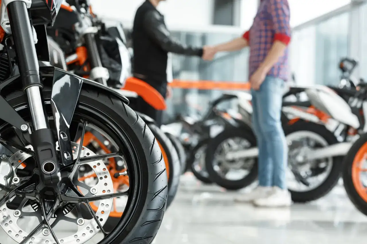 Dwóch mężczyzn ściska sobie dłonie przy motocyklach, finalizując transakcję. W tle widać rzędy maszyn, a na pierwszym planie szczegół koła. To może być moment zakupu, gdzie omawiany jest podatek od zakupu motocykla.
