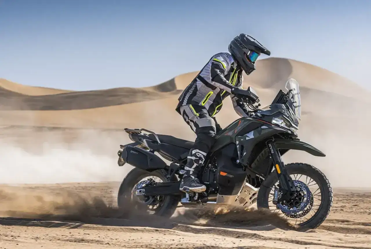 Motocyklista w pełnym rynsztunku pędzi na motocyklu KTM Adventure 790 przez pustynię, wzbijając tumany piasku.