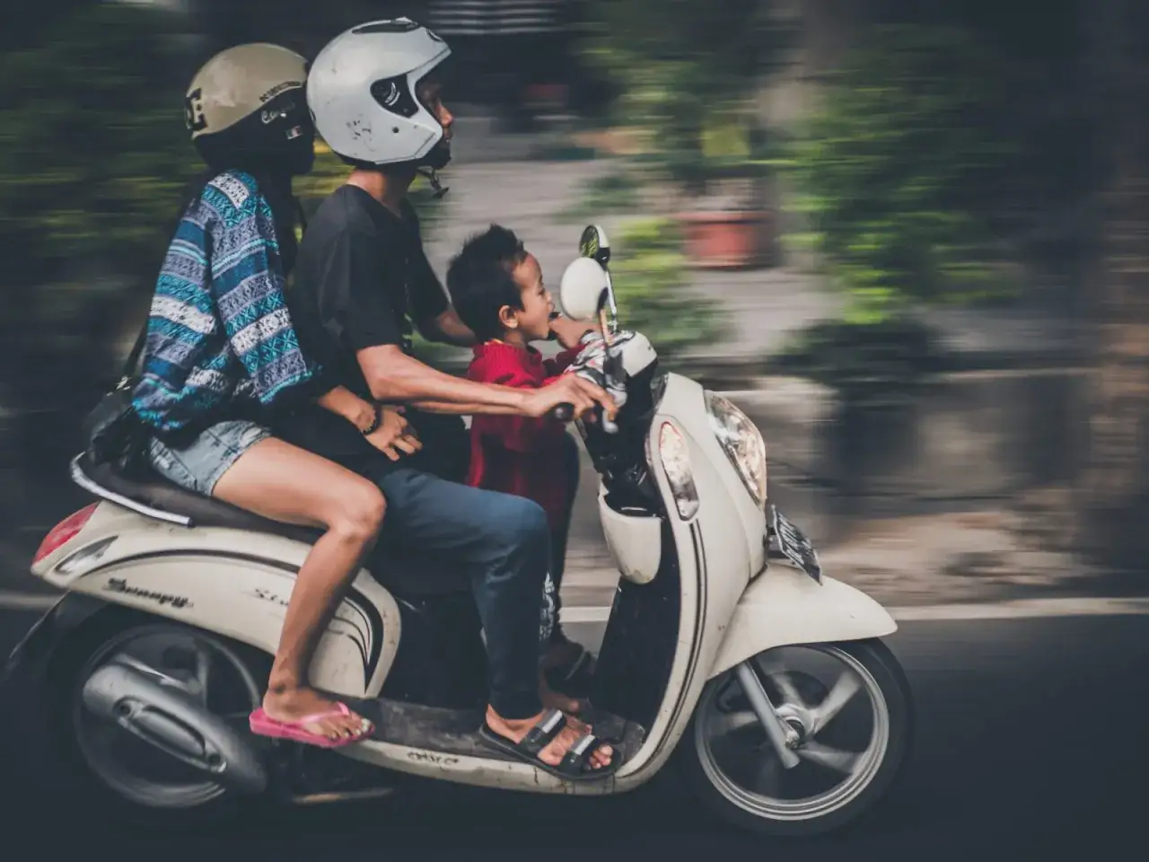 Rodzina na motocyklu, ojciec z dzieckiem z przodu, matka z tyłu, wszyscy w kaskach, pędzą ulicą.