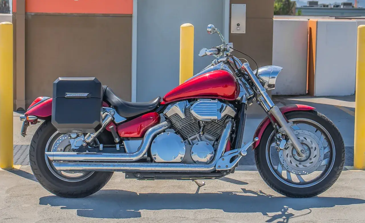Czerwony motocykl Honda VTX 1300 z czarnymi kuframi, zaparkowany przy żółtym słupku.
