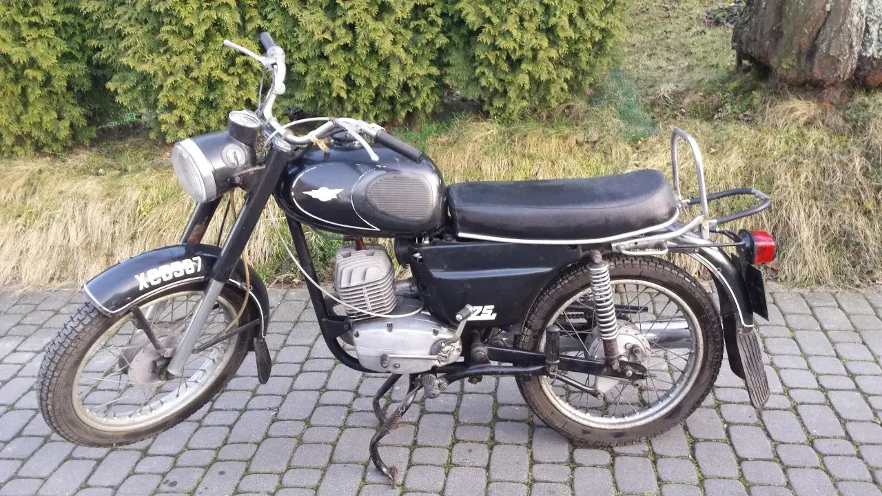 Czarny motocykl WSK 175, z widocznymi danymi technicznymi na silniku i błotniku. Stoi na chodniku z kostki brukowej, w tle krzewy i trawa.