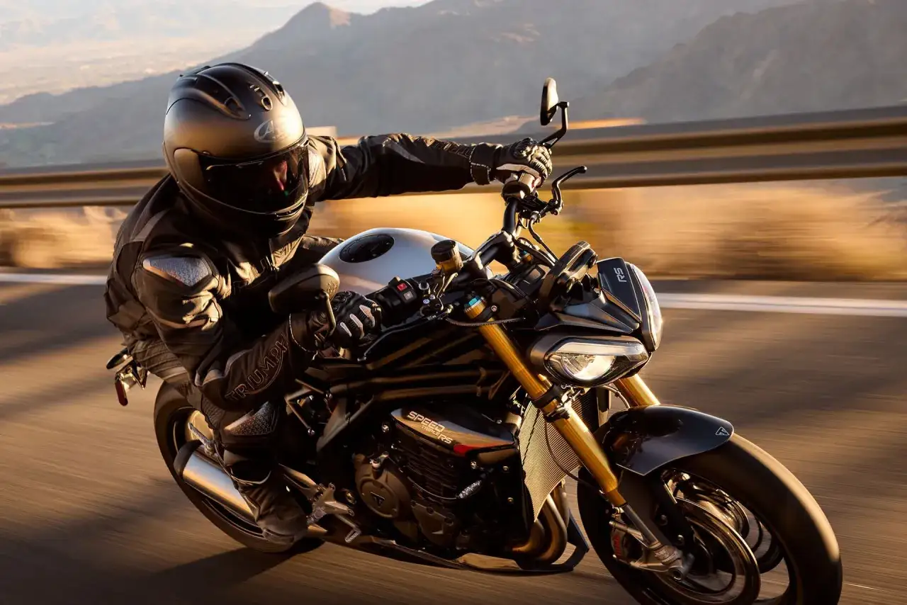 Motocyklista w kasku i kombinezonie pędzi na motocyklu Triumph Speed Triple RS po malowniczej drodze.