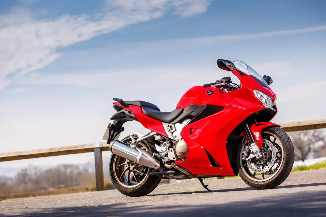 Czerwona Honda VFR 800 zaparkowana przy drewnianym płocie, na tle błękitnego nieba z chmurami.