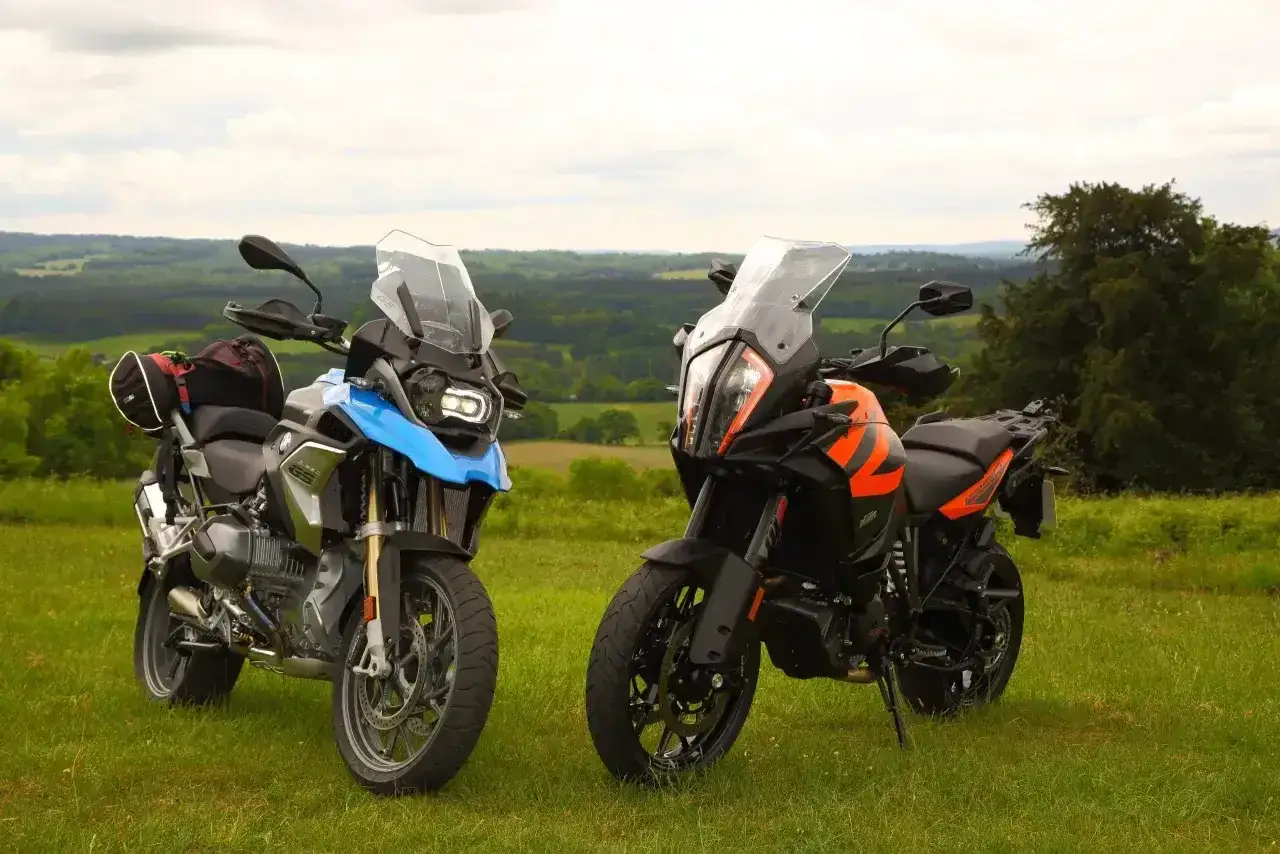 Dwa motocykle turystyczne, jeden niebieski, drugi pomarańczowy KTM 1290 Super Adventure, stoją na trawie z widokiem na zielone wzgórza.