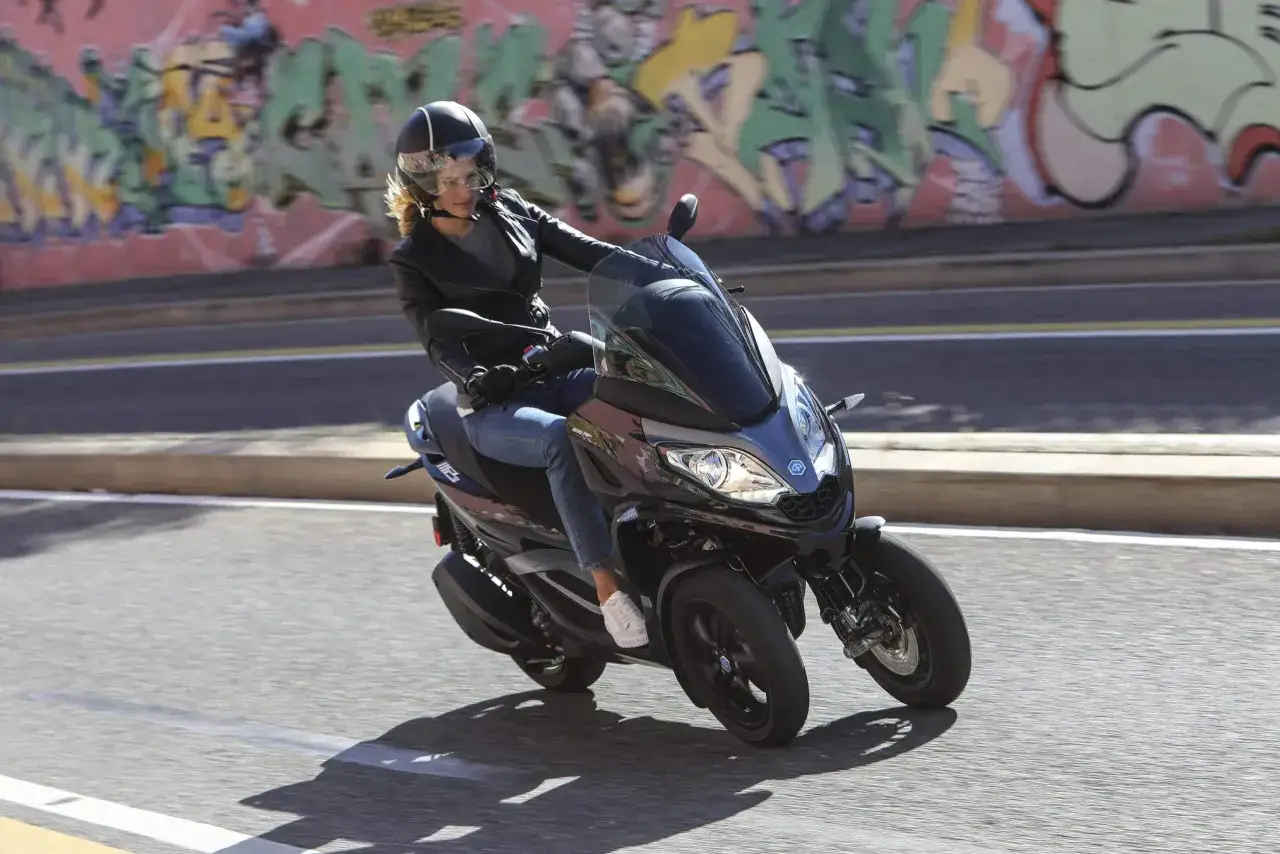Kobieta w kasku i skórzanej kurtce jedzie na trójkołowym skuterze Piaggio MP3 300 HPE. Tło stanowi kolorowy mural.