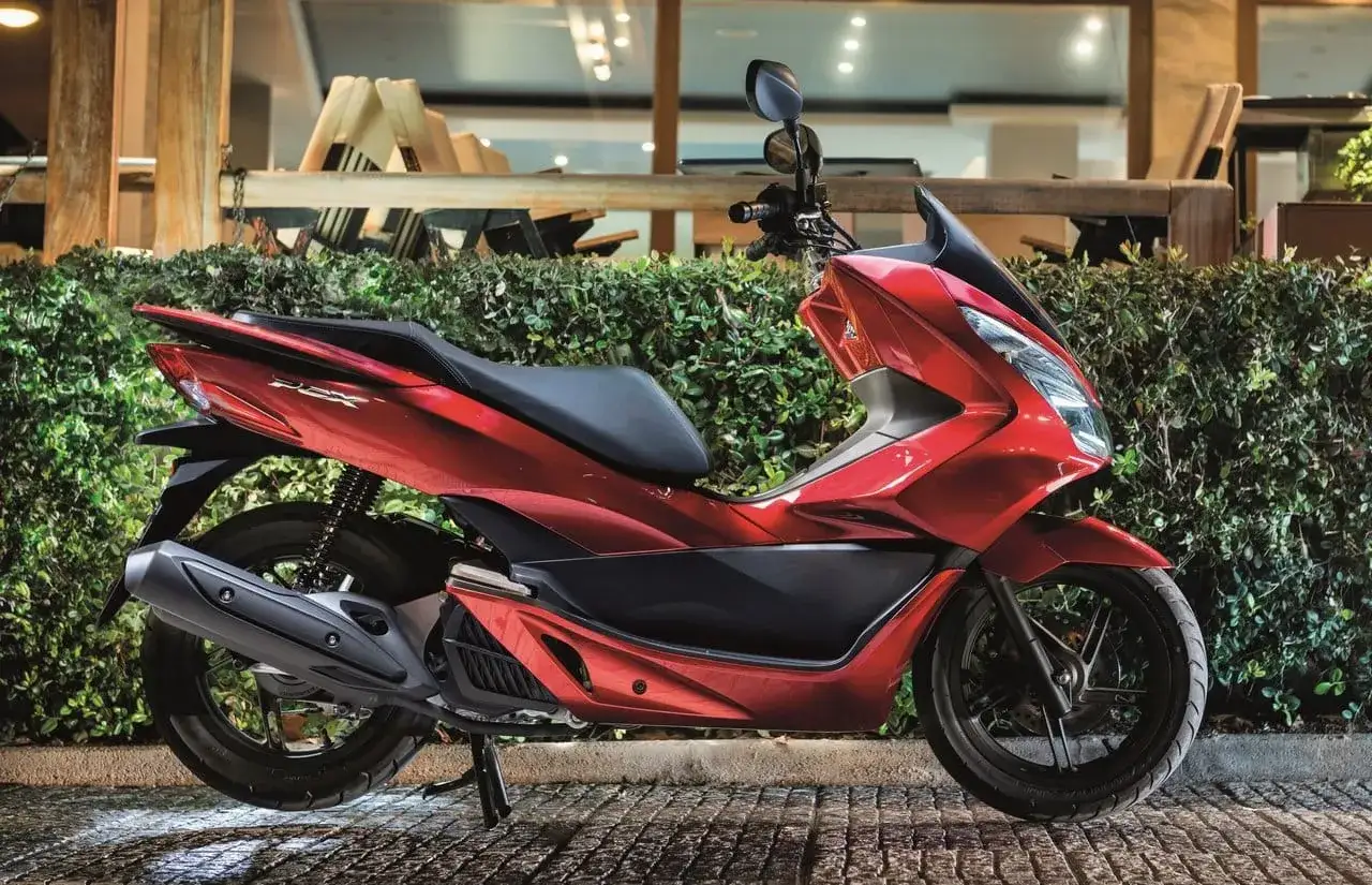 Czerwony skuter Honda PCX, jeden z najbardziej bezawaryjnych skuterów 125, zaparkowany przed zielonym żywopłotem.