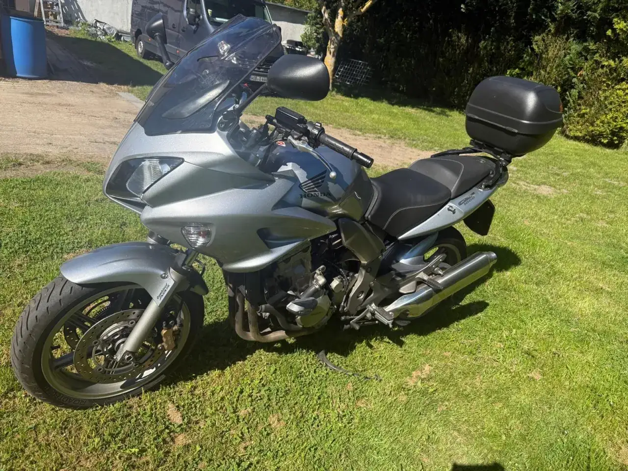 Srebrny motocykl Honda CBF 1000 z czarnym kufrem na bagaż, zaparkowany na trawie.