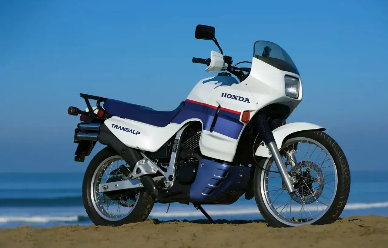 Honda Transalp 650 na plaży, gotowa na przygodę. Biało-niebieski motocykl z czerwonym paskiem.