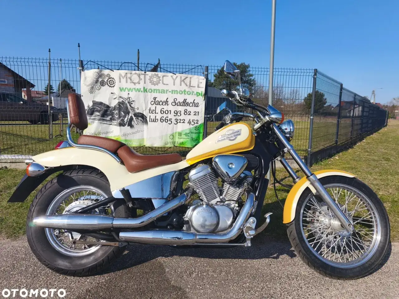 Żółto-kremowy motocykl Honda Shadow 500 z brązowym siedzeniem i bagażnikiem, zaparkowany przed banerem z ofertą motocykli.