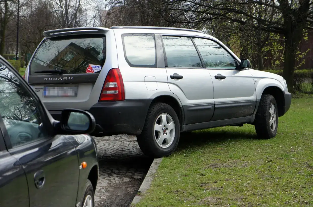 Srebrny Subaru Forester parkuje na trawniku, częściowo na chodniku. Obok stoi ciemny samochód.