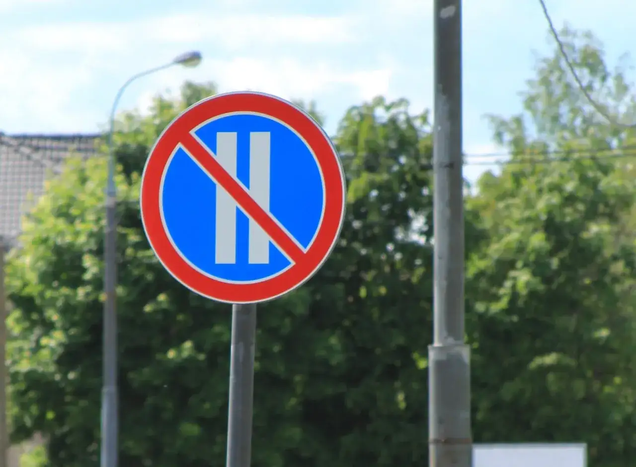 Znak B37 zakazujący postoju. Dwa pionowe białe paski na niebieskim tle, przekreślone czerwoną linią.