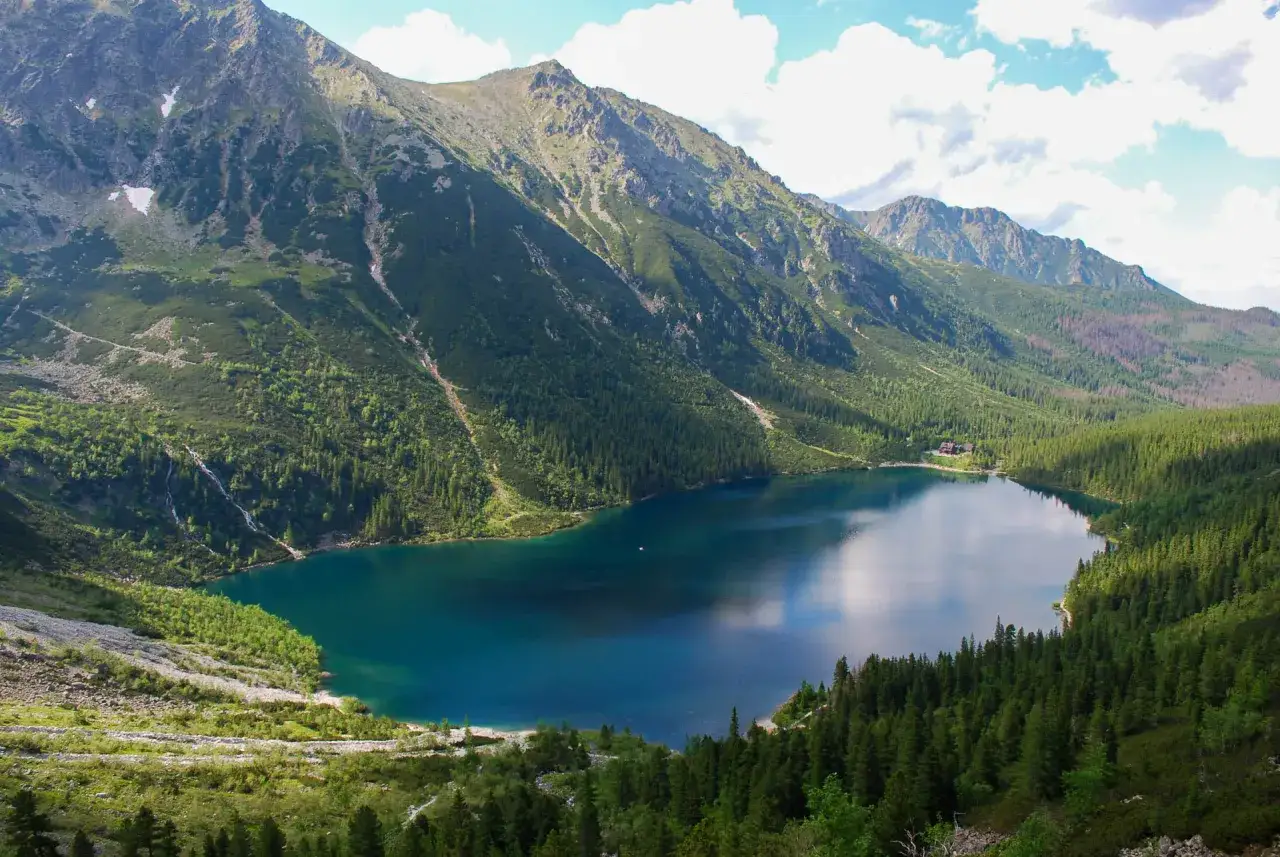 Malowniczy widok na Morskie Oko, z górami i lasami otaczającymi turkusową taflę wody. Droga na Morskie Oko wije się wśród zieleni.
