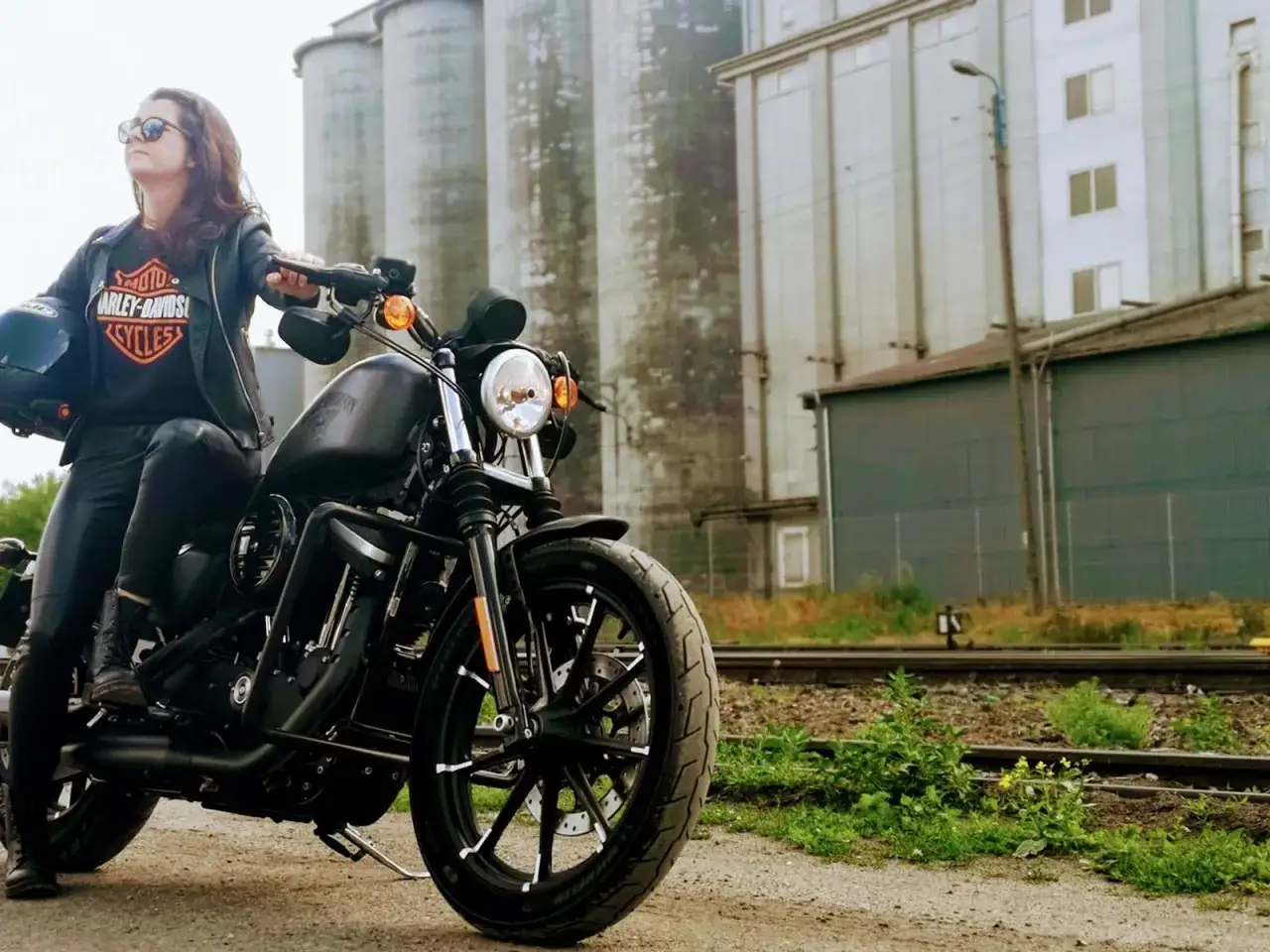 Kobieta w skórzanej kurtce i goglach siedzi na czarnym Harley-Davidson Sportster 883, gotowa do jazdy.