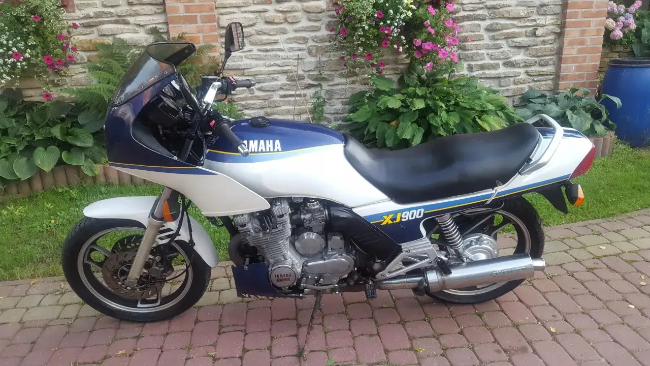Klasyczny motocykl Yamaha XJ 900 w kolorze białym z granatowymi i żółtymi akcentami, zaparkowany na brukowanym podjeździe.