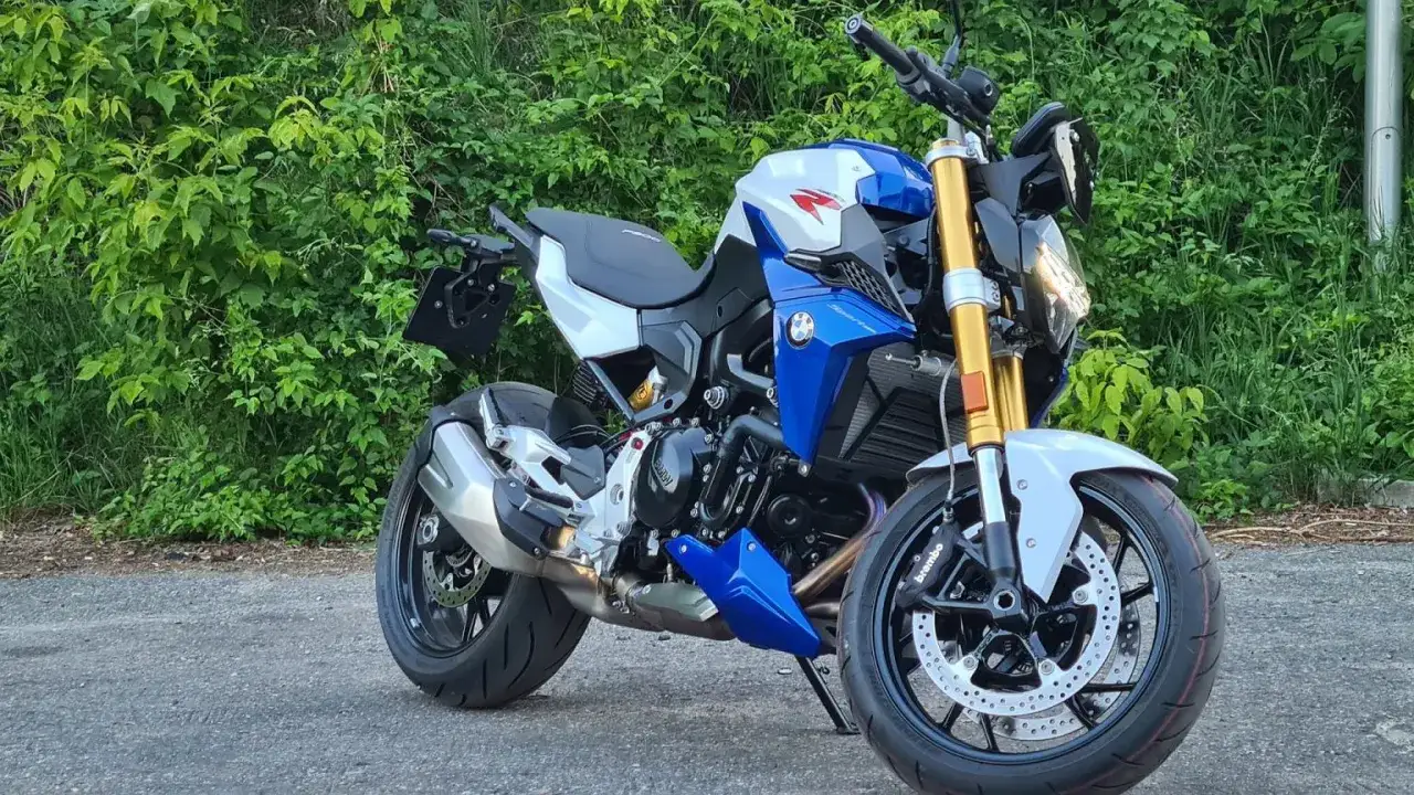 Błękitno-biały motocykl BMW F900R z błyszczącymi złotymi lagami, gotowy do jazdy pośród zieleni.