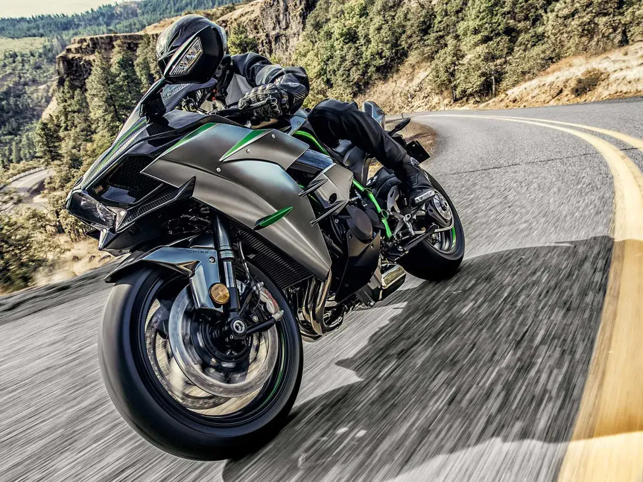 Motocyklista na Kawasaki Ninja H2 pokonuje zakręt na górskiej drodze, otoczony zielenią i skałami.