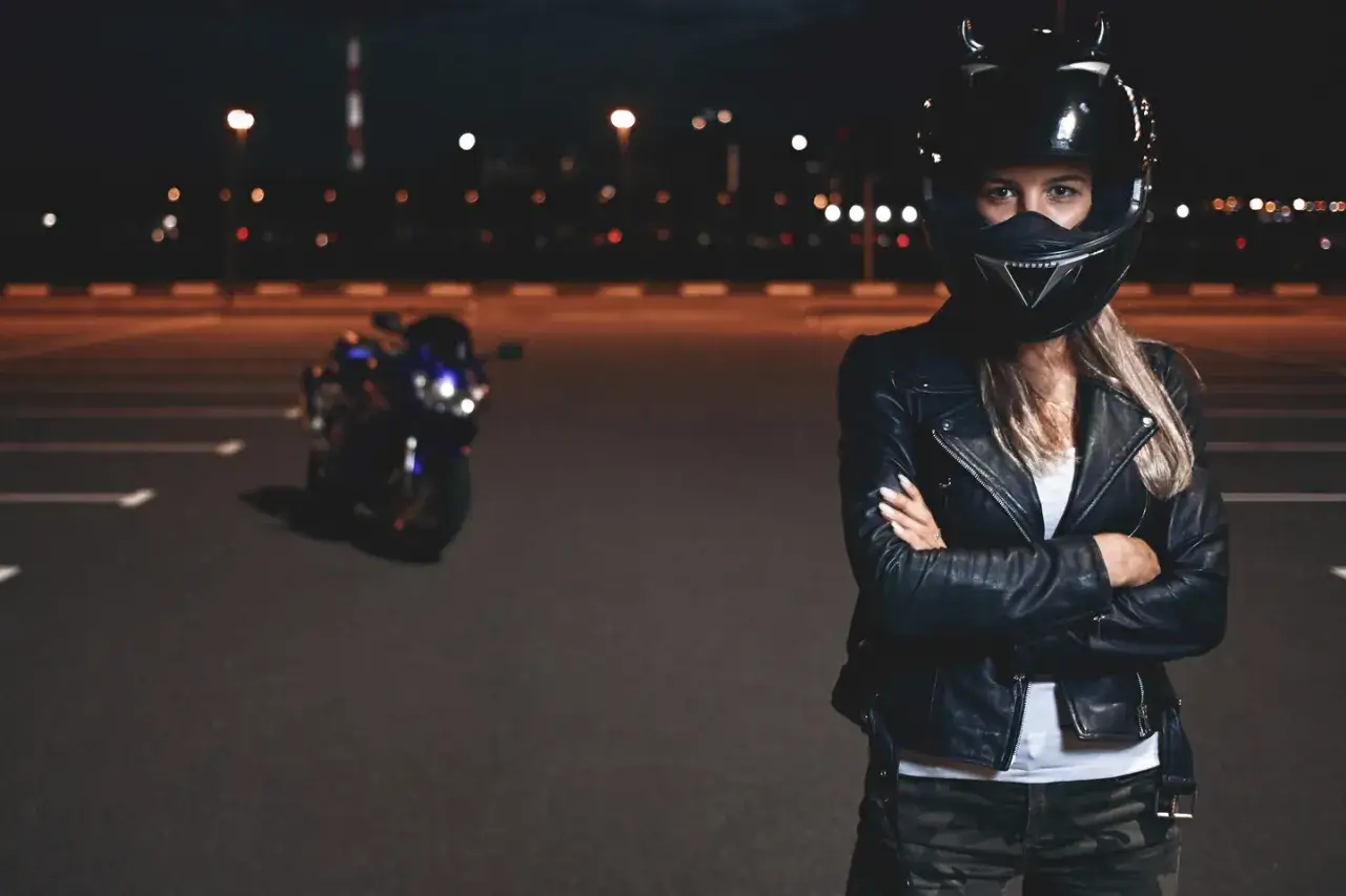 Kobieta w kasku motocyklowym i skórzanej kurtce stoi na tle nocnego parkingu z motocyklem. W oddali widać światła drogowe.