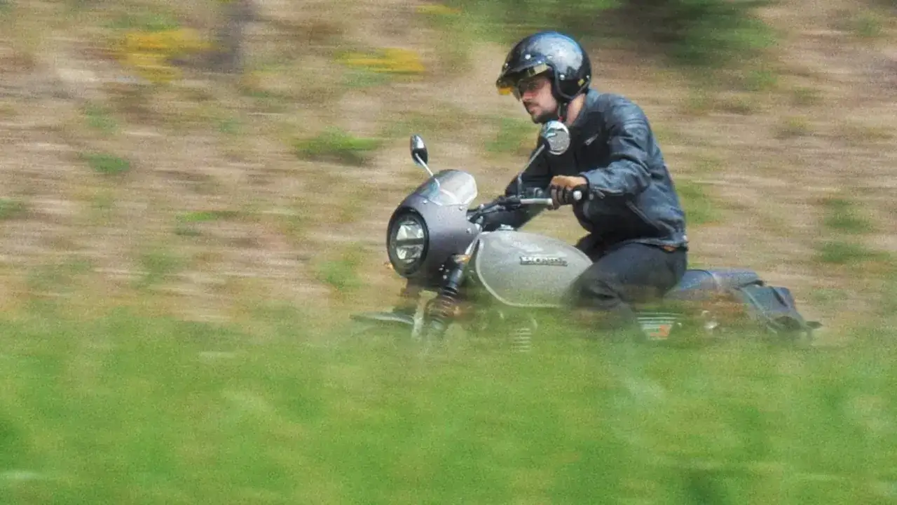 Mężczyzna w kasku i skórzanej kurtce jedzie motocyklem Honda GB350S przez zielone pola.