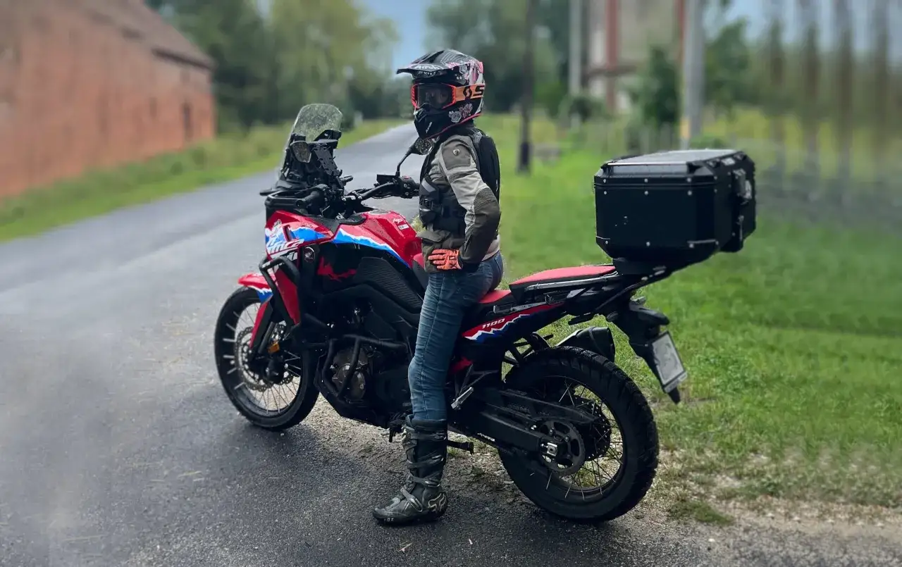 Kobieta w kasku i odzieży motocyklowej stoi obok czerwonego motocykla adventure z czarnym kufrem.
