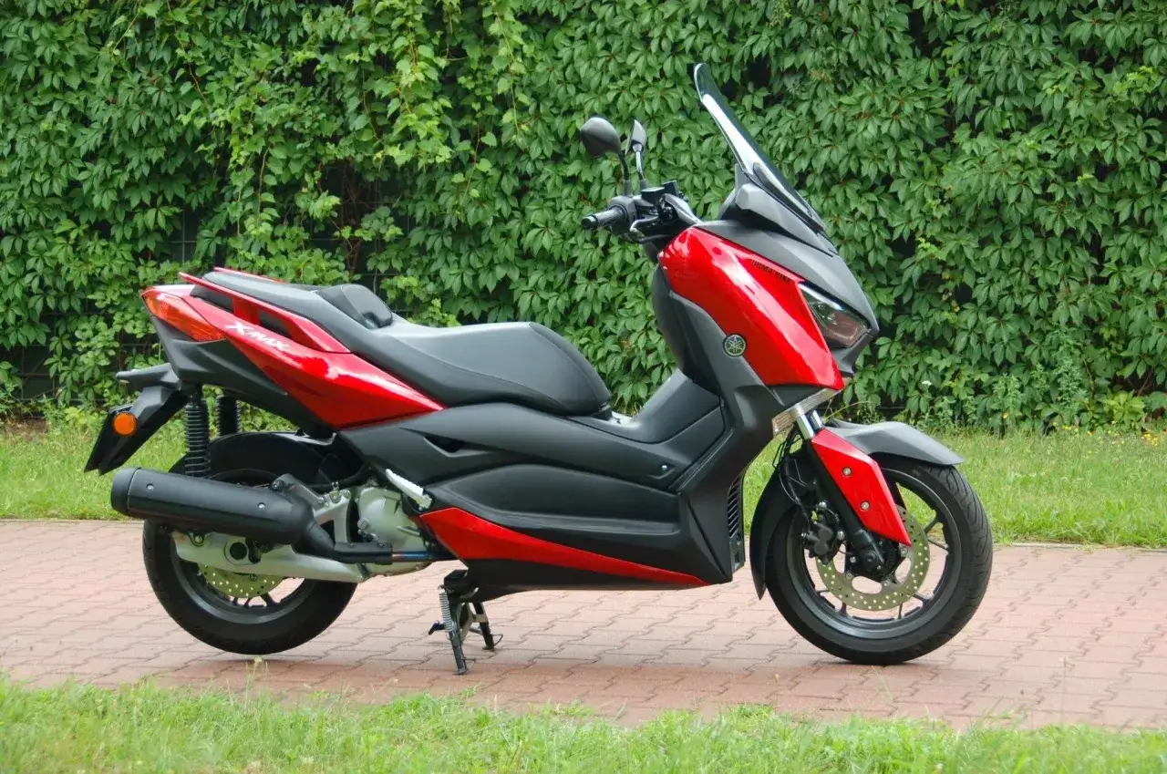 Czerwony skuter Yamaha X-MAX 125, gotowy do osiągnięcia swojej prędkości maksymalnej na tle zielonego żywopłotu.