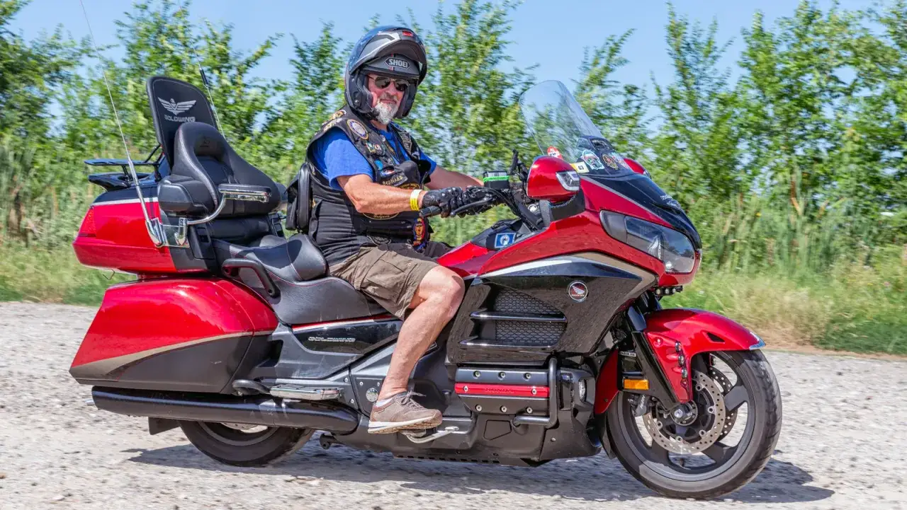 Mężczyzna w kasku jedzie na czerwonym motocyklu honda turystyk. W tle zielone drzewa i błękitne niebo.