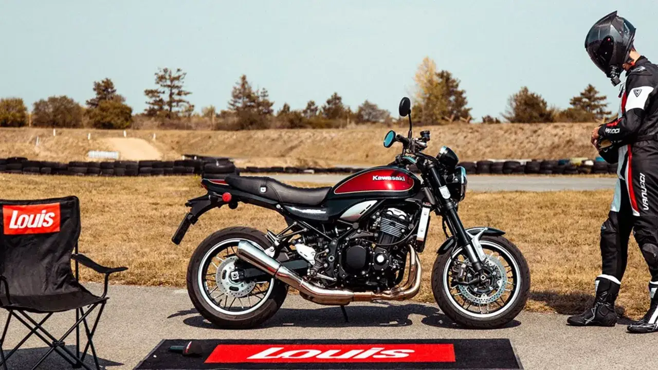 Klasyczny motocykl Kawasaki Z900RS w kolorze czarno-czerwonym stoi na torze wyścigowym obok krzesła z logo Louis.
