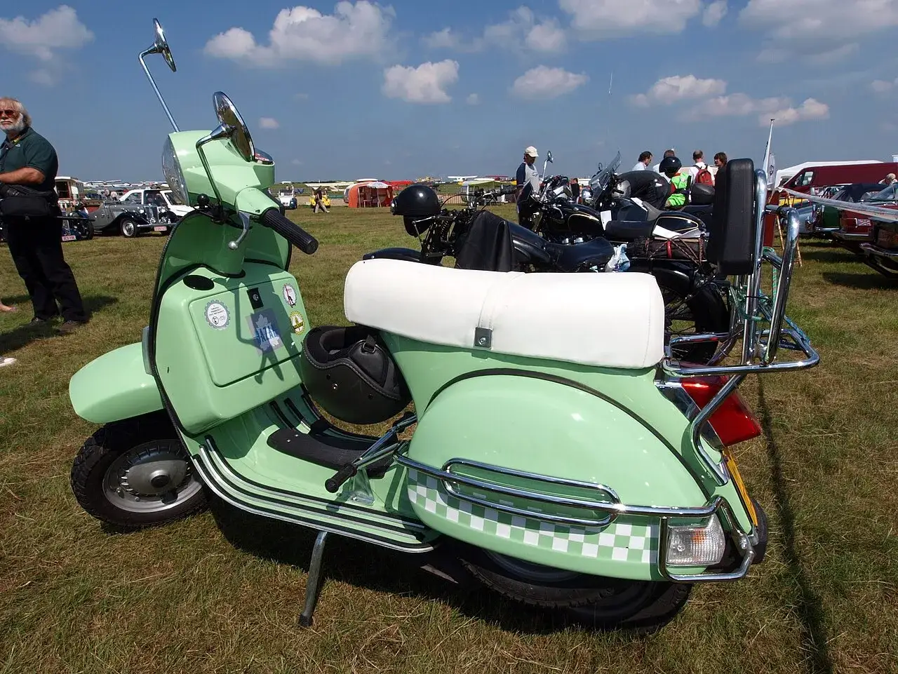 Jasnozielona Vespa z białym siedzeniem i czarnym kaskiem, gotowa na przejażdżkę. W tle inne klasyki i ludzie. #lml