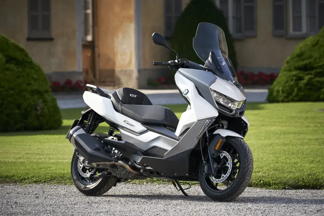 Biały skuter BMW C400GT z szarymi akcentami, zaparkowany na żwirowej ścieżce przed budynkiem z zielonym ogrodem.